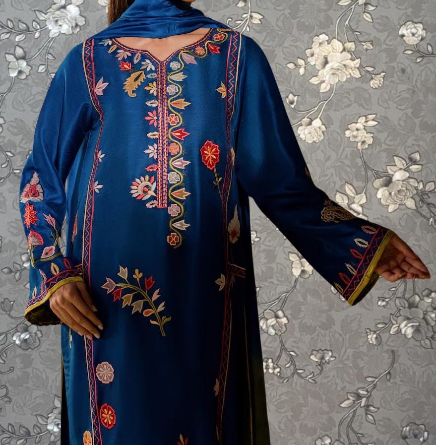 A person wearing a blue dress with intricate embroidered patterns on the sleeves and back, posing against a gray floral wallpaper background.