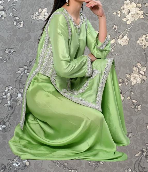 A woman wearing a long, green dress with silver embroidery is sitting on the floor against a gray floral background.