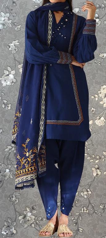 A woman wearing a blue dress with intricate embroidery and a matching blue shawl stands against a gray floral background.