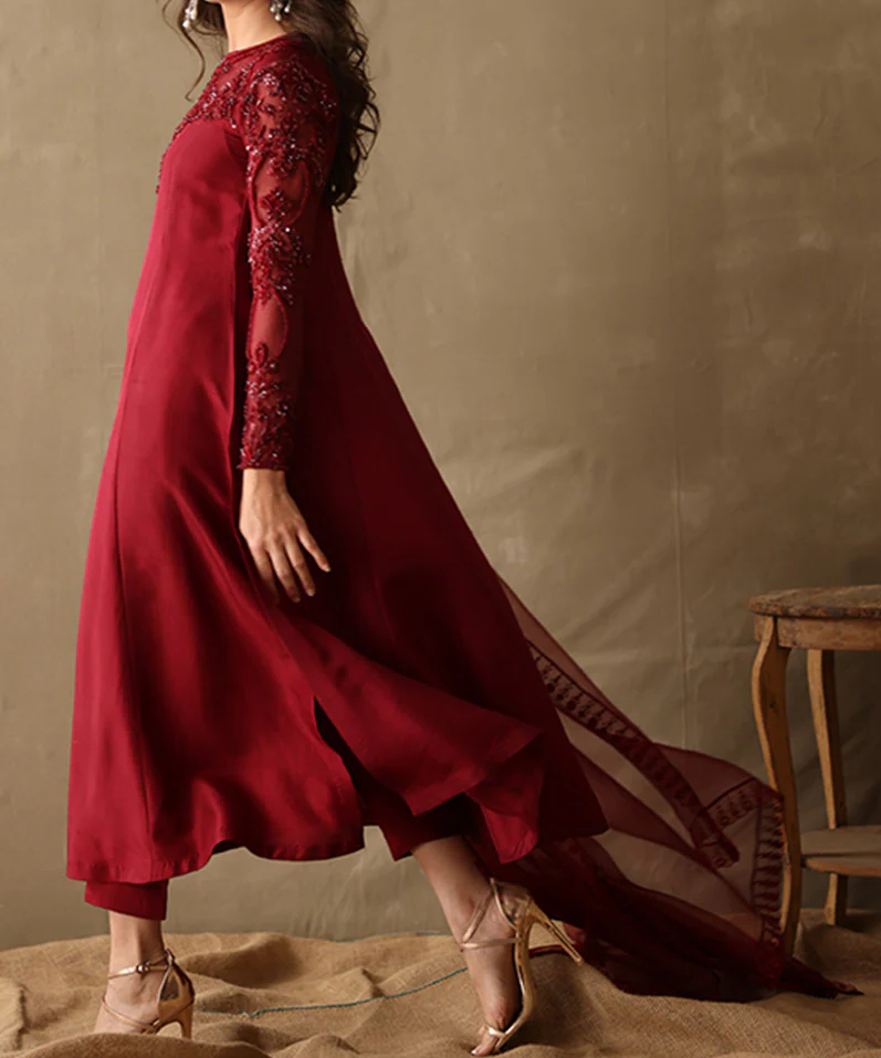 A woman wearing a long, red dress with intricate embroidery and a flowing train walks in front of a wooden chair.