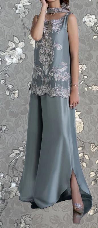 A woman wearing a gray dress with intricate embroidery and a high slit, posing against a gray floral background.