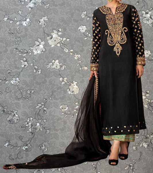 A woman wearing a black dress with gold embroidery and a long black shawl, standing against a gray floral background.
