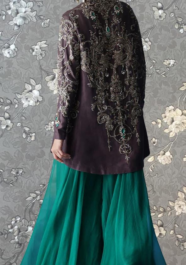 a person wearing a long, dark purple jacket with intricate embroidery and a matching green skirt. The person is standing against a gray floral background.