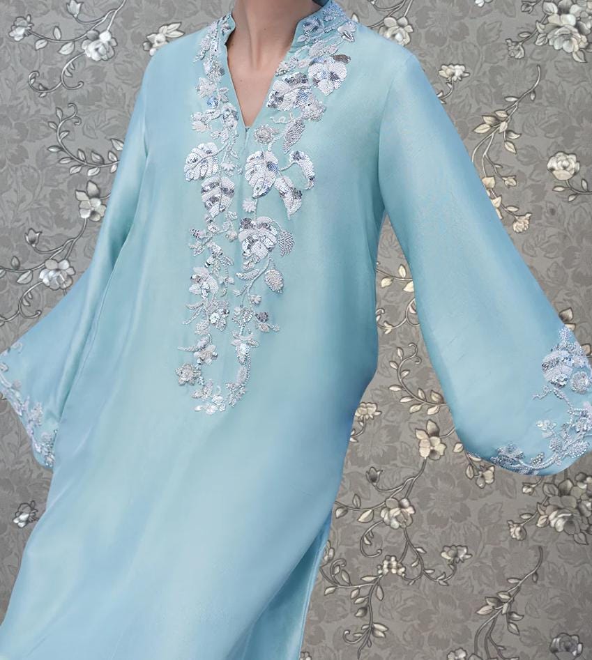 A close-up of a light blue dress with intricate floral embroidery on the neckline and sleeves, set against a gray floral background.