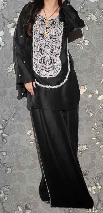 A woman wearing a black dress with a white lace pattern on the front, standing against a gray floral background.