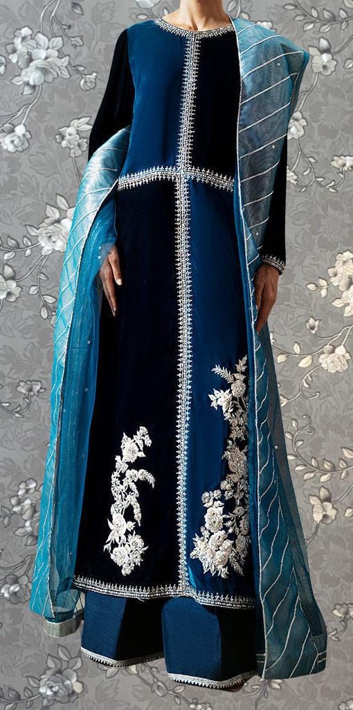 A person wearing a blue velvet dress with white floral embroidery, standing against a gray floral background.