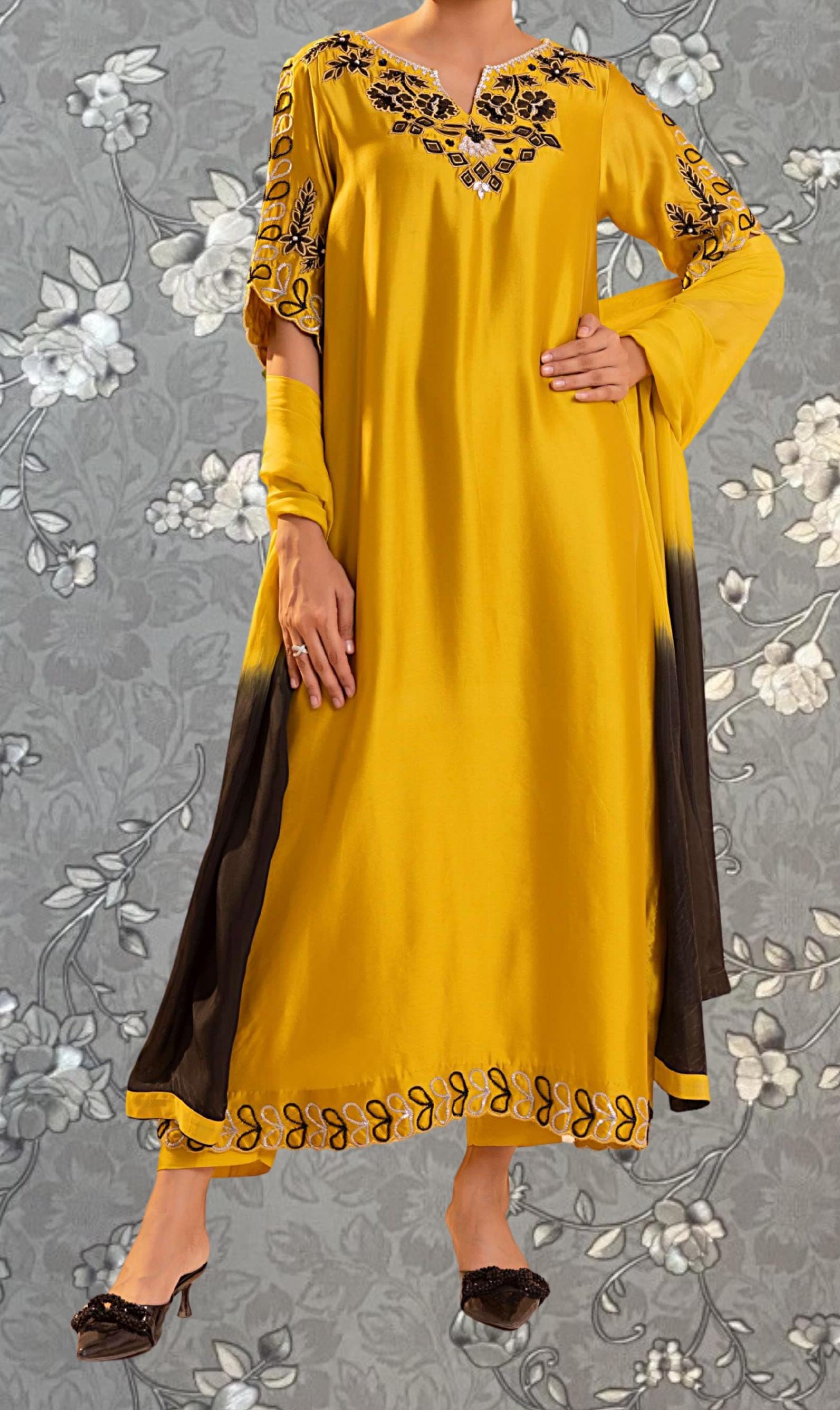 A woman wearing a long yellow dress with black embroidered details, posing against a gray floral background.