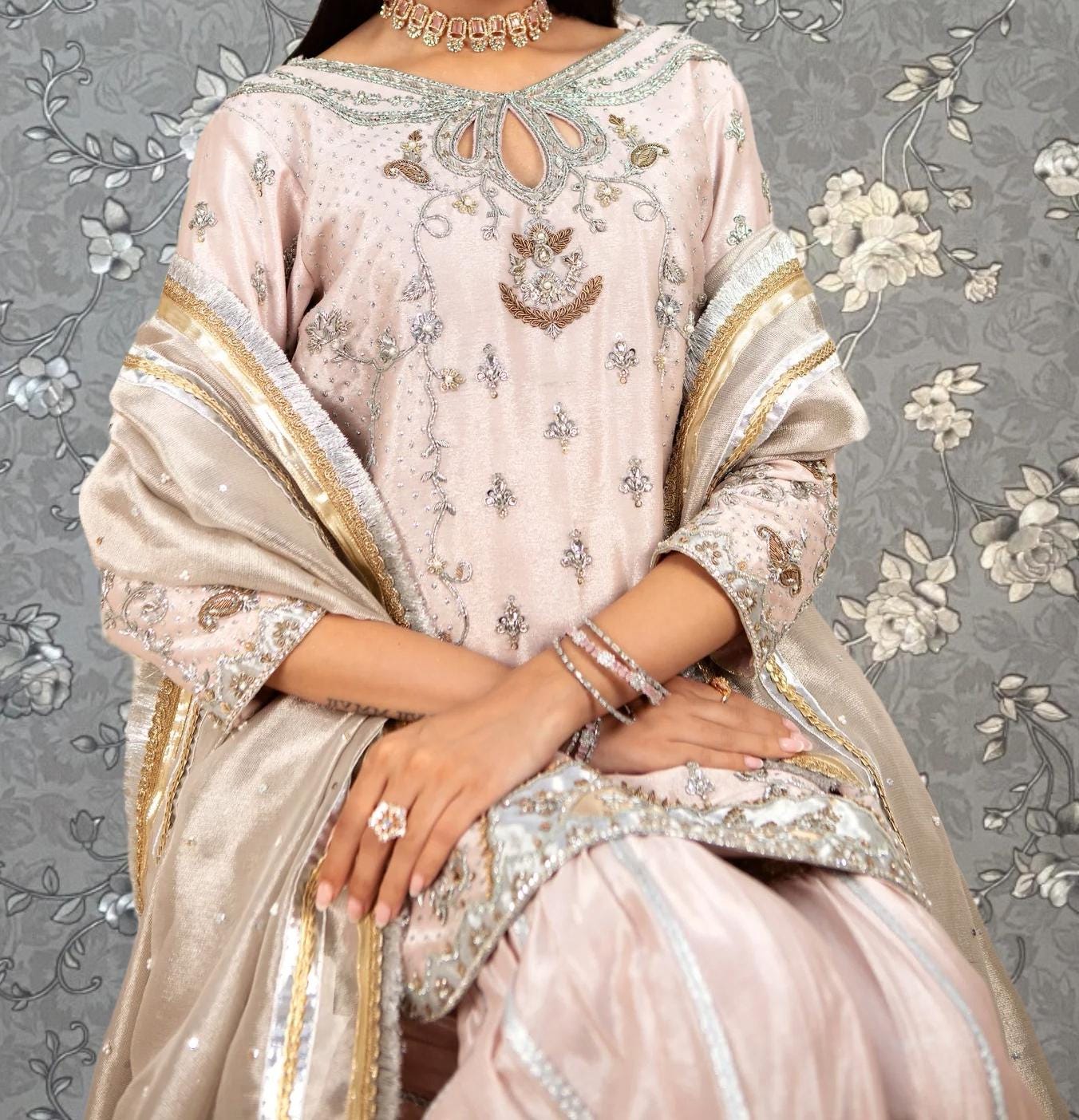 A woman wearing a pink and gold dress with intricate embroidery and jewelry poses against a gray floral background.