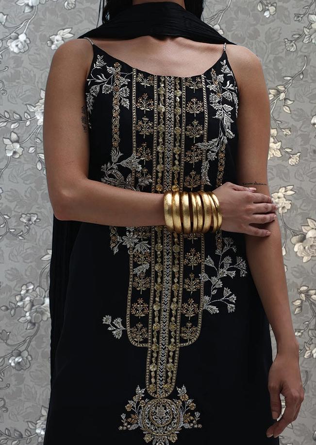 A woman wearing a black dress with gold embroidery and gold bracelets stands against a floral background.