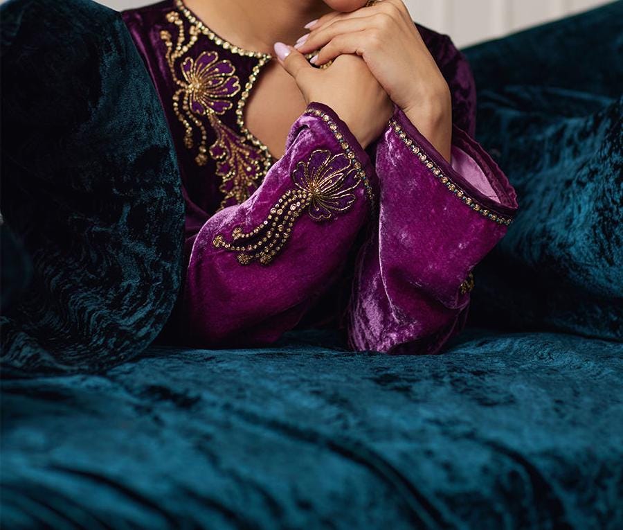 A close-up of a person wearing a purple velvet dress with gold embroidery, resting their hands on their lap.