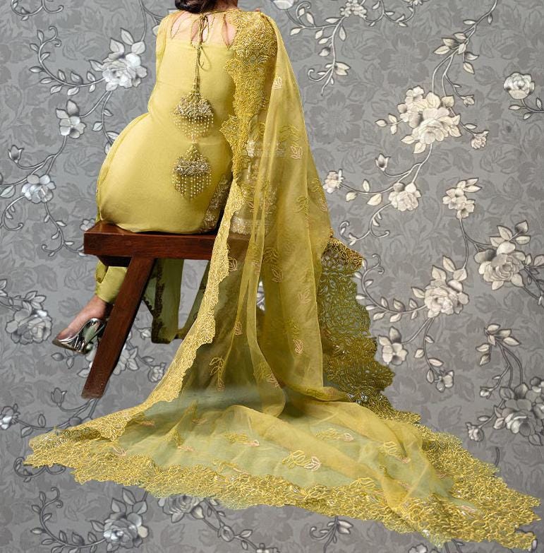 A woman wearing a yellow dress with a long, flowing train sits on a wooden stool against a gray floral background.
