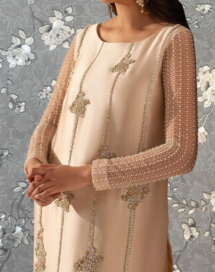 A woman wearing a beige dress with intricate embroidery and beaded sleeves stands against a floral background.