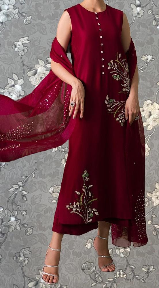 A woman wearing a red dress with floral embroidery and a matching red shawl stands against a gray floral background.