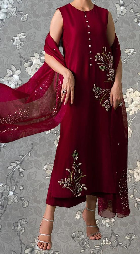 A woman wearing a red dress with floral embroidery and a matching red shawl stands against a gray floral background.