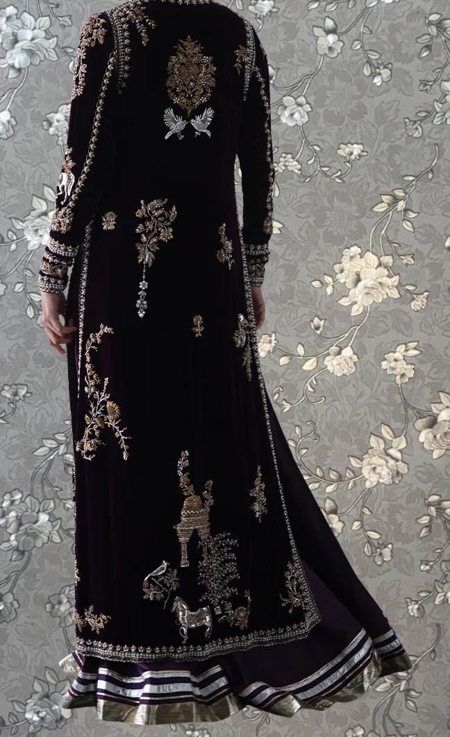 a person wearing a long, dark purple velvet dress with intricate embroidered designs. The dress has a high collar and long sleeves, giving it a formal and elegant appearance. The background features a floral pattern, which complements the overall design of the dress.