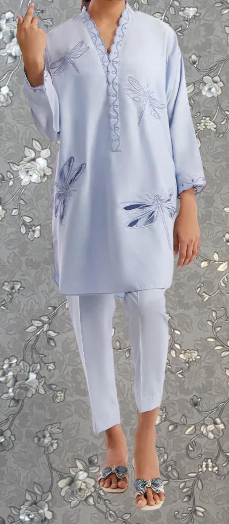 A person wearing a light blue dress with blue dragonfly designs on it, standing against a gray floral background.