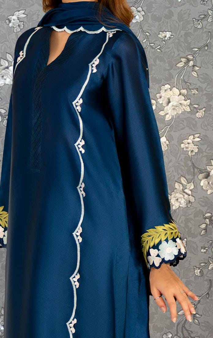 A woman wearing a blue dress with white floral embroidery on the sleeves and collar stands against a gray floral background.
