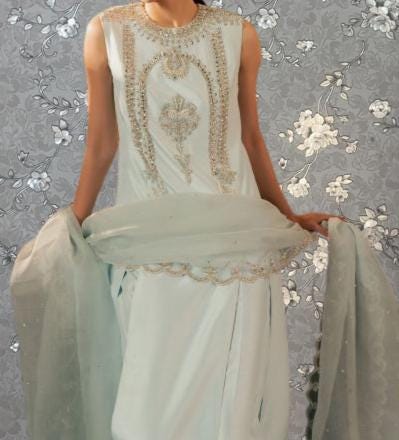 A woman wearing a white dress with intricate embroidery stands against a gray floral background.