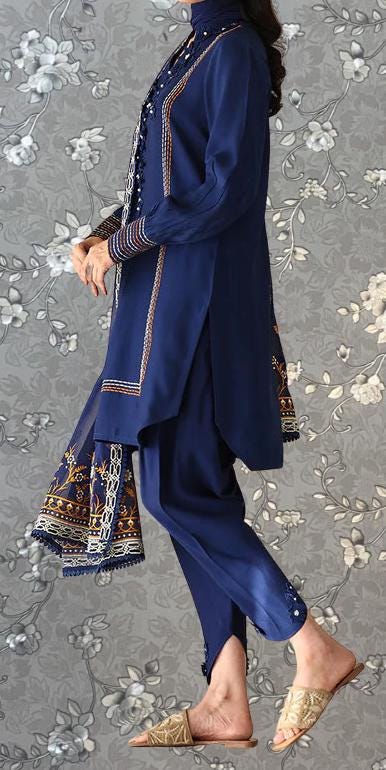 A person wearing a blue dress with intricate embroidery and gold accents, standing against a gray floral background.