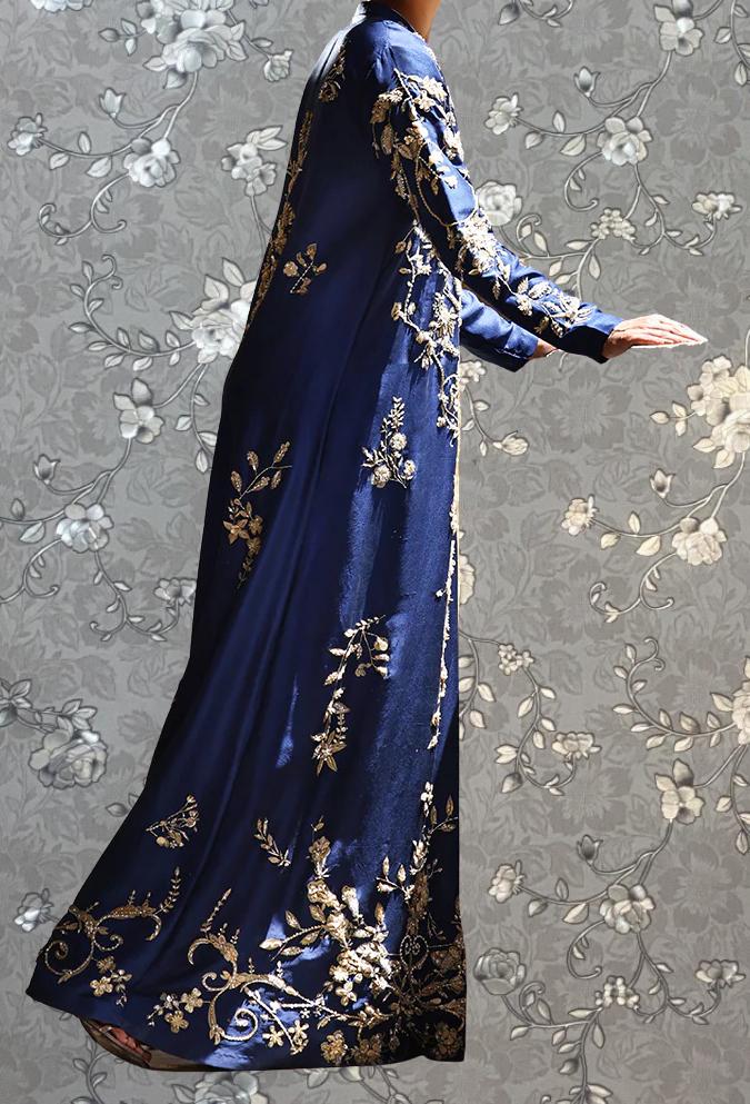 A person wearing a long, blue dress with gold embroidery stands against a gray floral background.