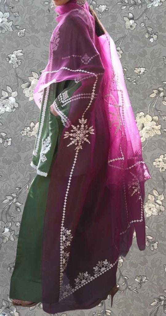 A person wearing a long, flowing dress with a pink and purple patterned fabric. The dress has a high neckline and a long train, and the person is standing against a gray floral background.