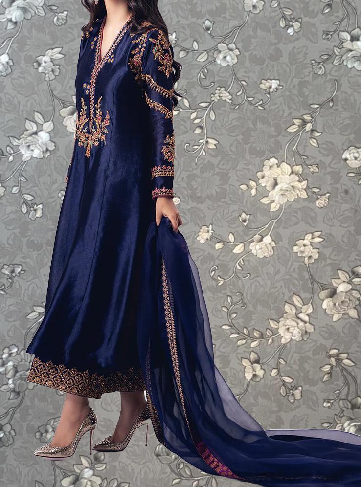 A woman wearing a long, blue dress with gold embroidery and high heels stands against a gray floral background.