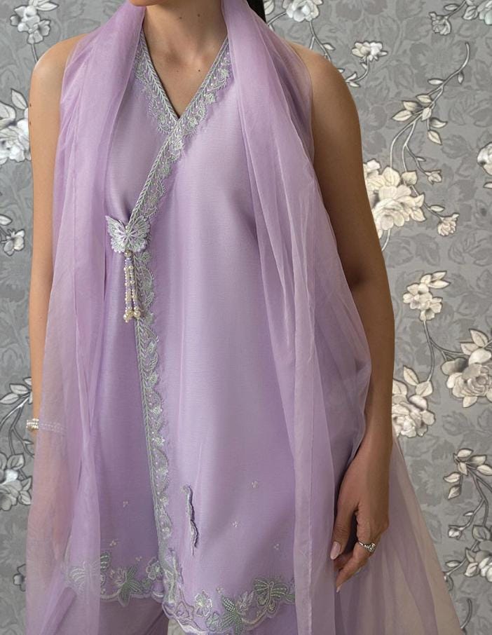 A woman wearing a lavender dress with a matching scarf stands against a gray floral background.