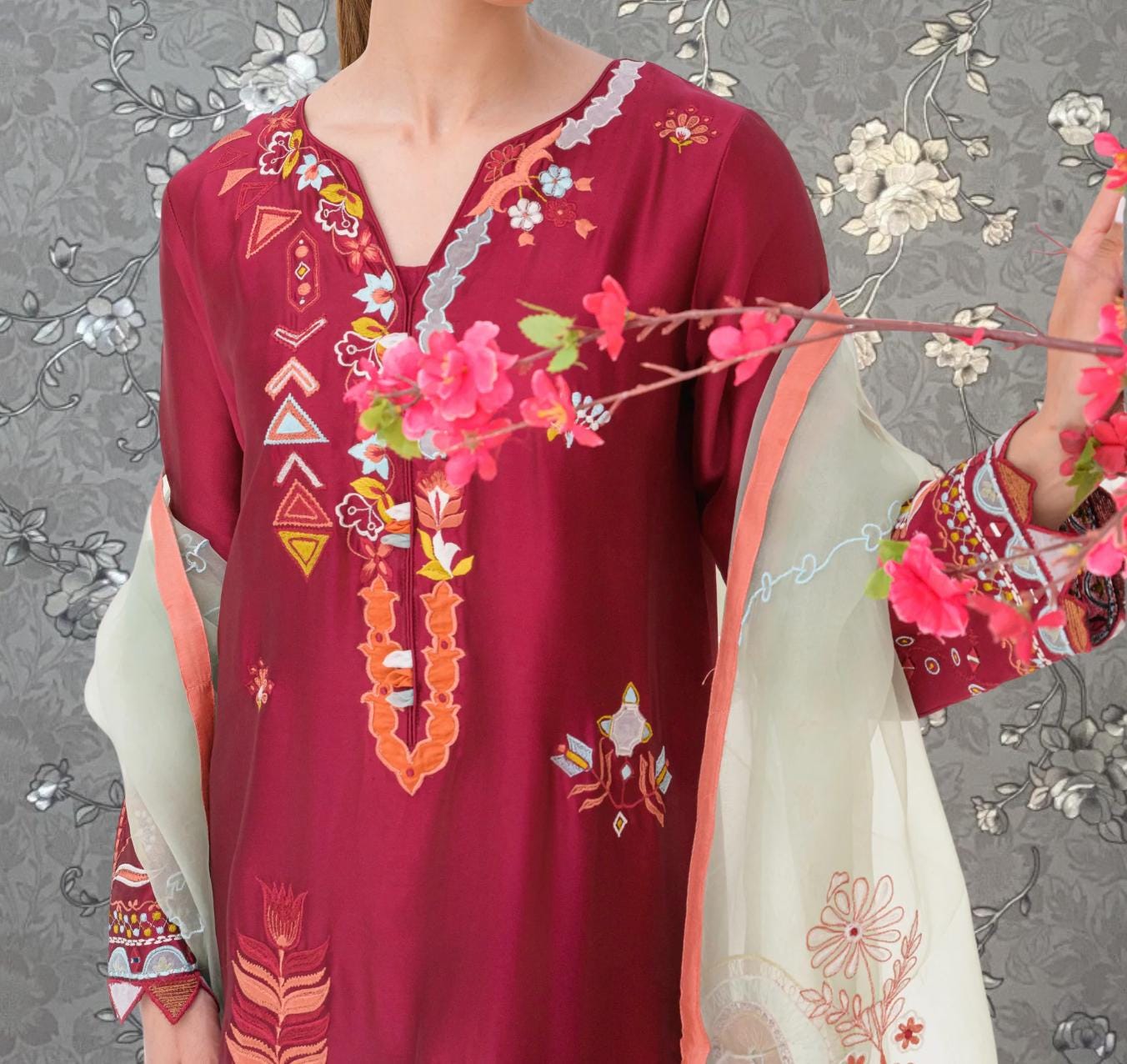 A woman wearing a red dress with intricate embroidery and floral designs, holding a branch with pink flowers.