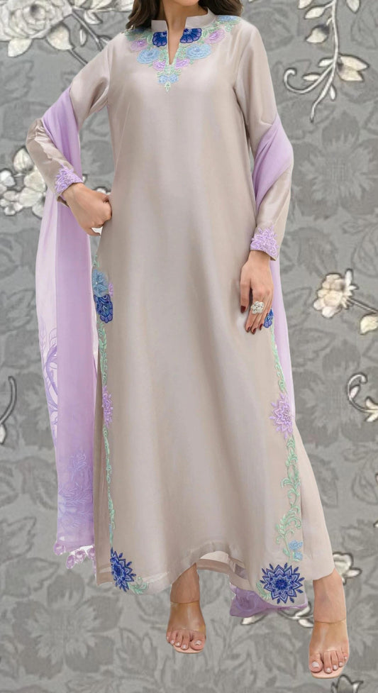 A woman wearing a long, light-colored dress with floral embroidery on the sleeves and hem. She is standing against a gray floral background.