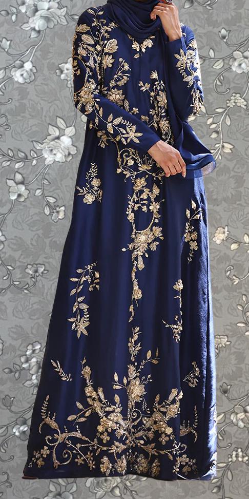 A woman wearing a long, blue dress with gold embroidery stands against a gray floral background.