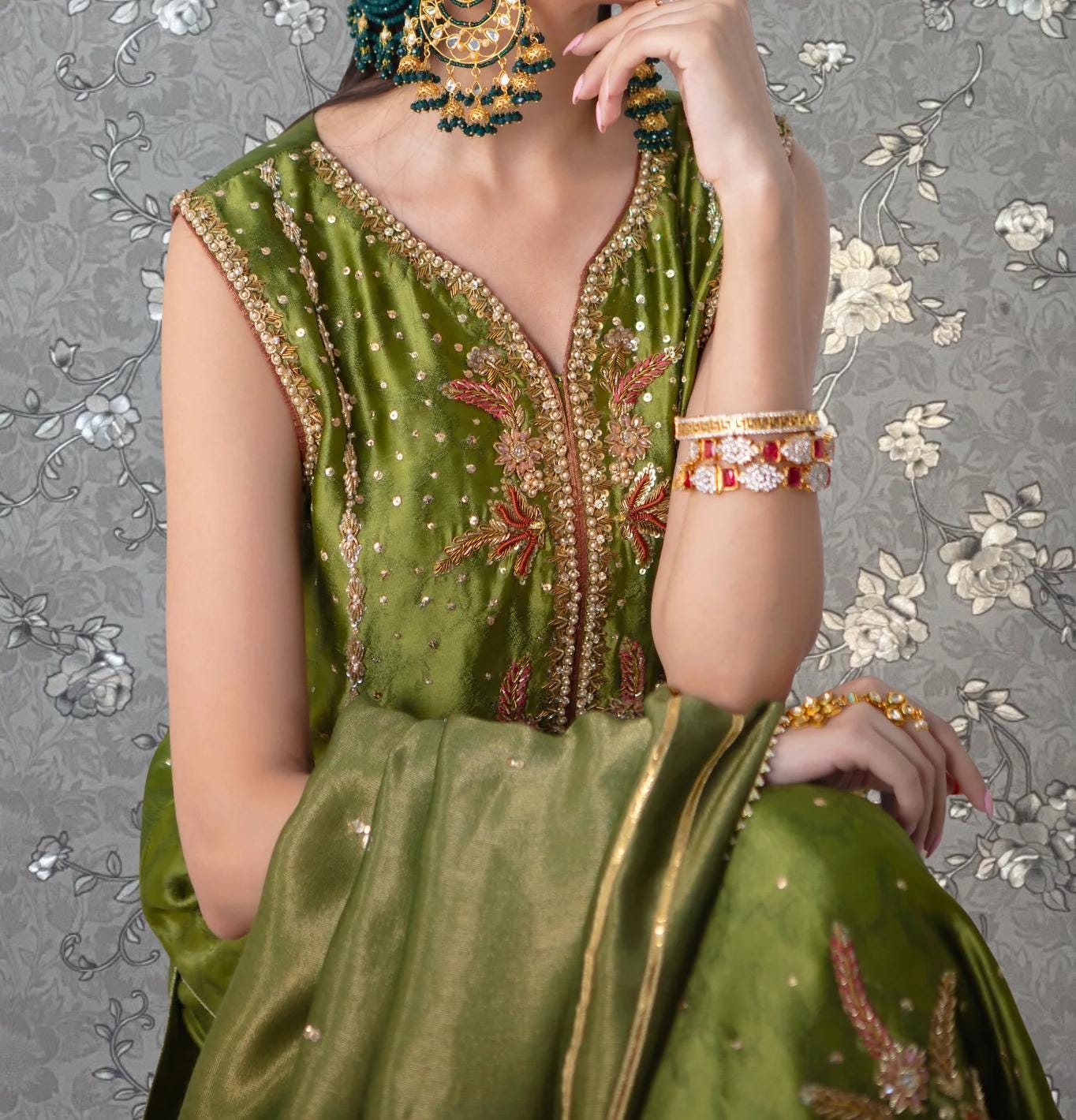 A woman wearing a green and gold dress with intricate embroidery and jewelry poses against a floral background.