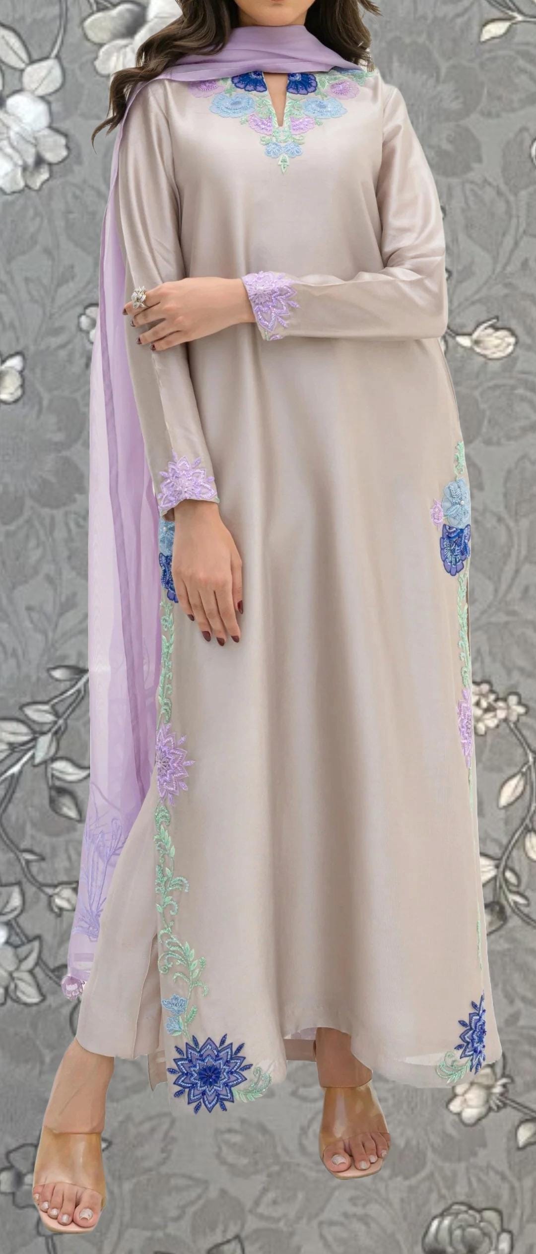 A woman wearing a long, light-colored dress with floral embroidery on the sleeves and hem. She is wearing a purple scarf and has her hands crossed.