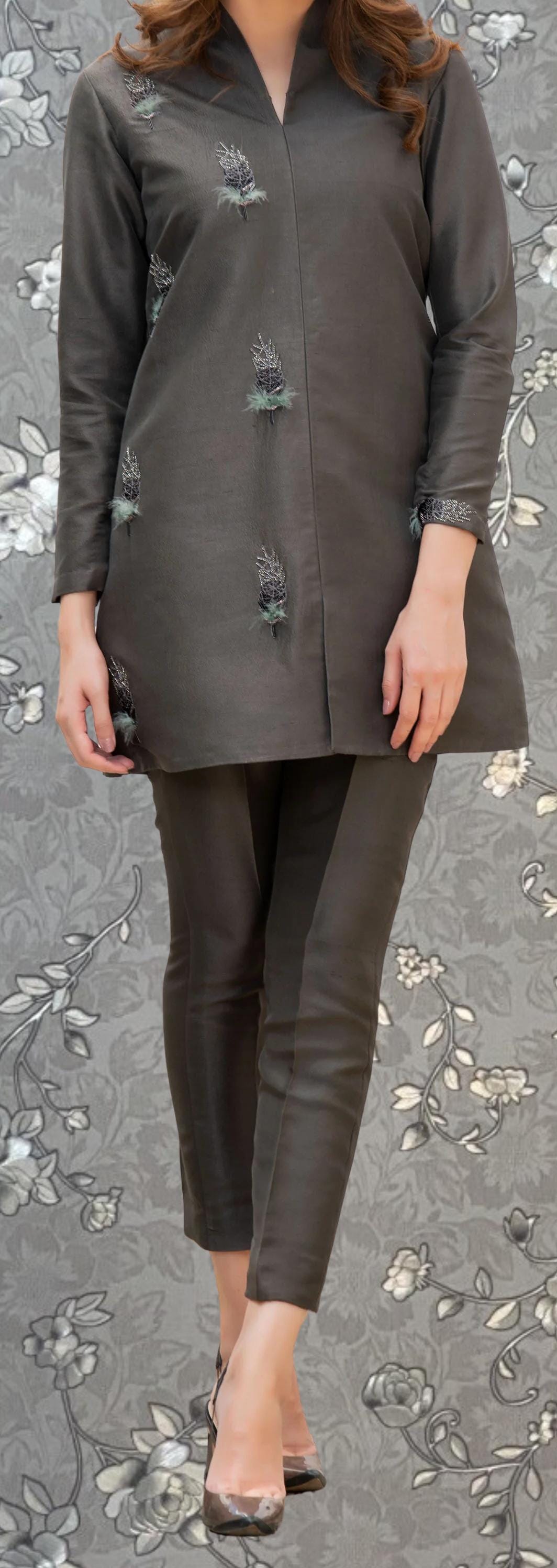 A woman wearing a gray dress with floral embroidery stands against a gray floral background.