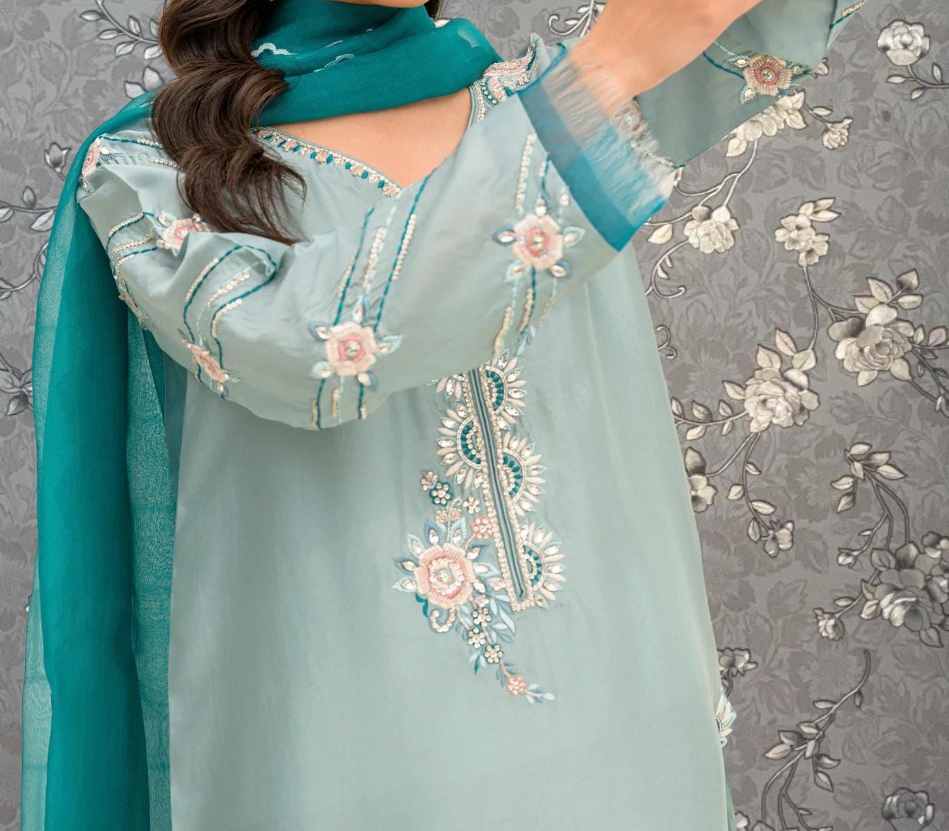 A woman wearing a light blue dress with intricate floral embroidery stands against a gray floral background.