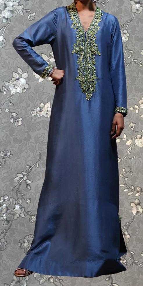 A person wearing a long, blue dress with a green and gold embroidered neckline and long sleeves, posing against a gray floral background.