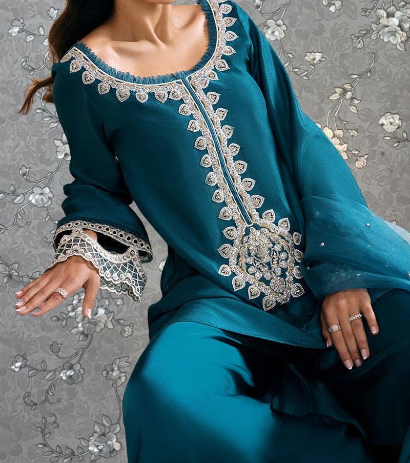 A woman wearing a teal blue dress with intricate embroidery and lace details, posing against a gray floral background.