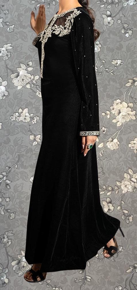 A woman wearing a long black dress with lace detailing and beaded sleeves stands against a gray floral background.