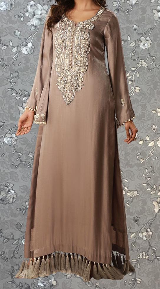 A woman wearing a long, beige dress with intricate embroidery and tassels on the bottom.