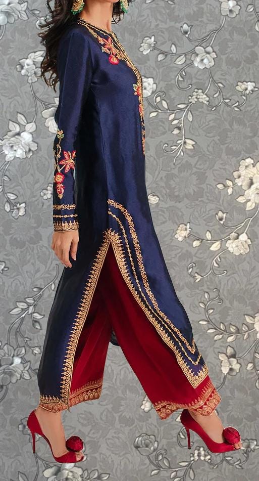 A woman wearing a long navy blue dress with gold embroidery and red pants, standing against a gray floral background.