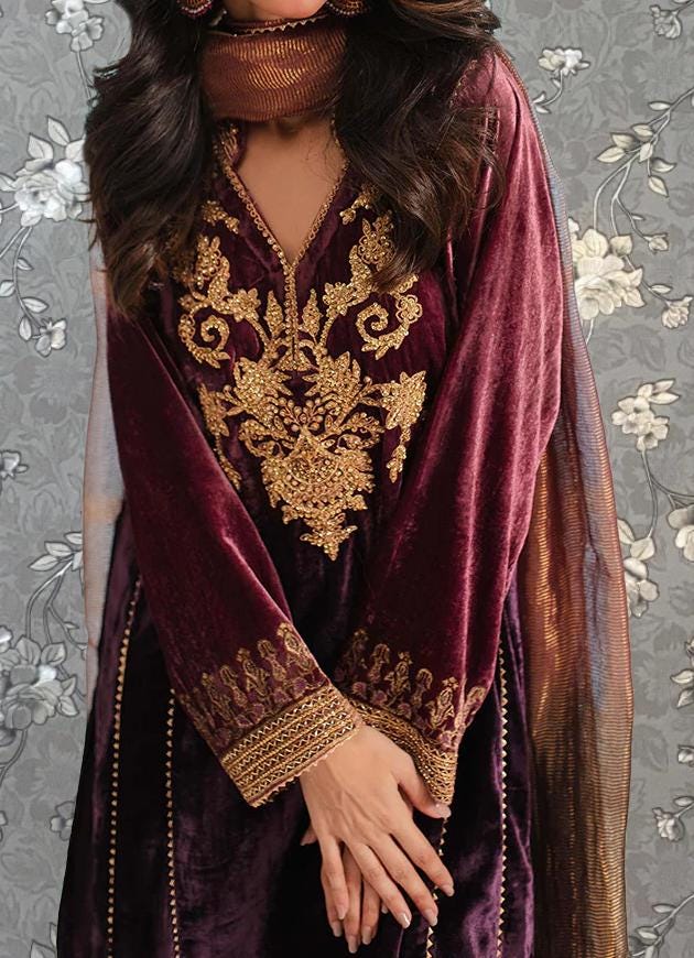 A woman wearing a long, burgundy velvet dress with gold embroidery stands against a gray floral background.