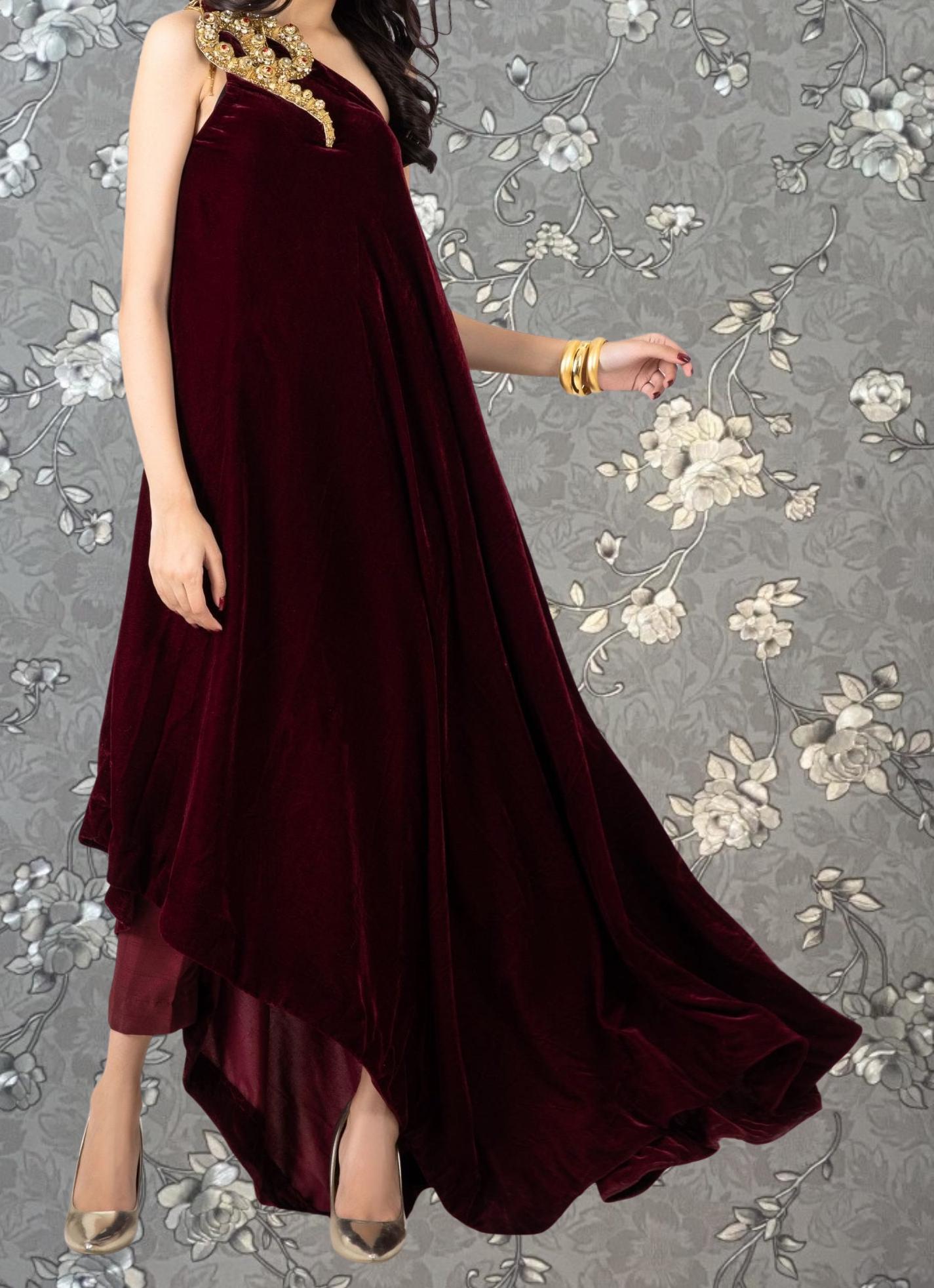 A woman wearing a long, burgundy velvet dress with a gold embellishment on the shoulder, posing against a gray floral background.