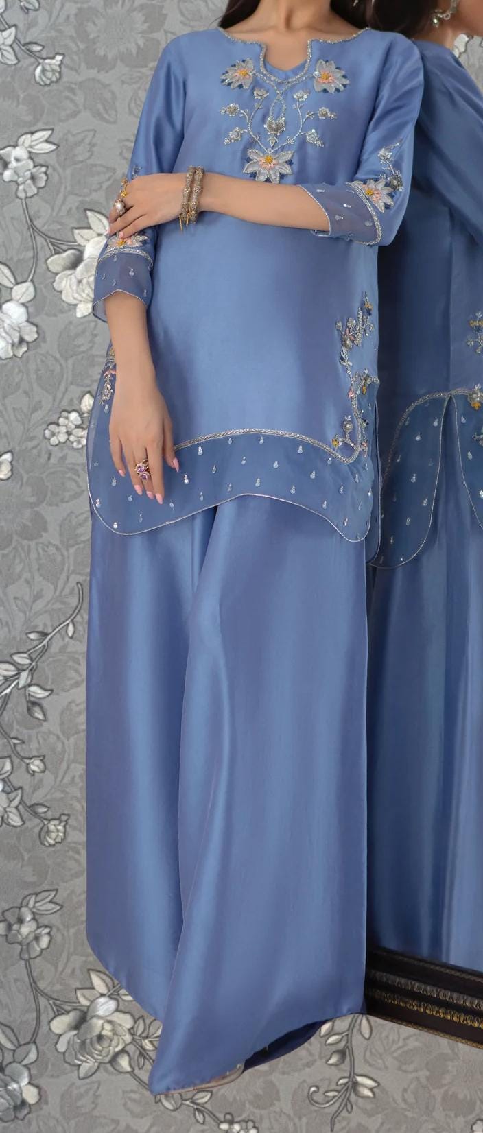 A woman wearing a blue dress with intricate embroidery and jewelry poses in front of a mirror.