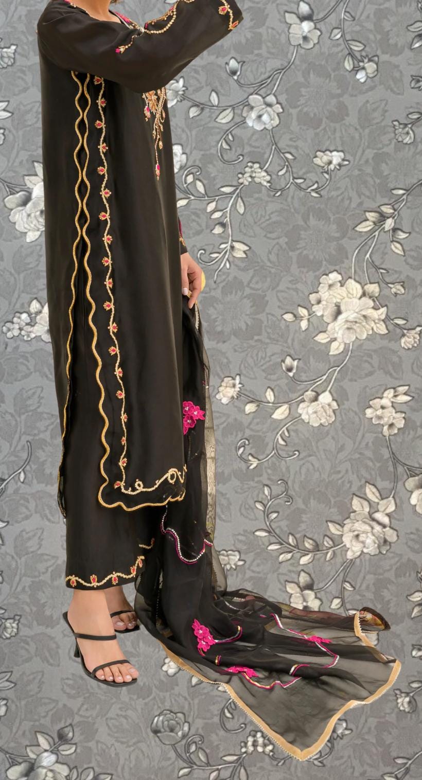 A person wearing a long black dress with gold trim and floral embroidery stands against a gray floral background.