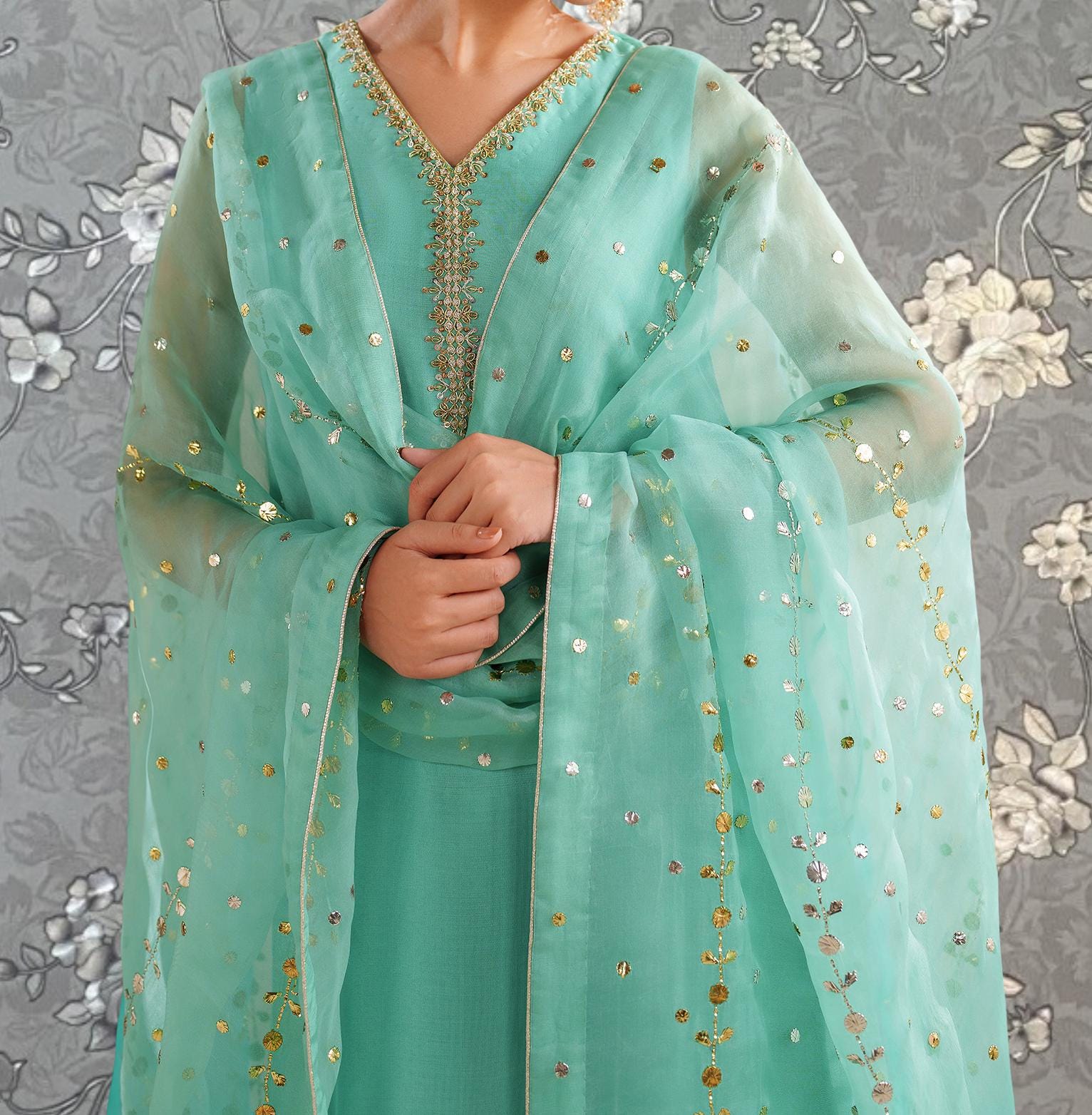 A person wearing a mint green dress with gold embroidery and a matching green shawl stands against a gray floral background.