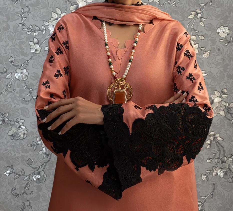 A person wearing a pink dress with black lace sleeves and a gold necklace stands against a gray floral background.