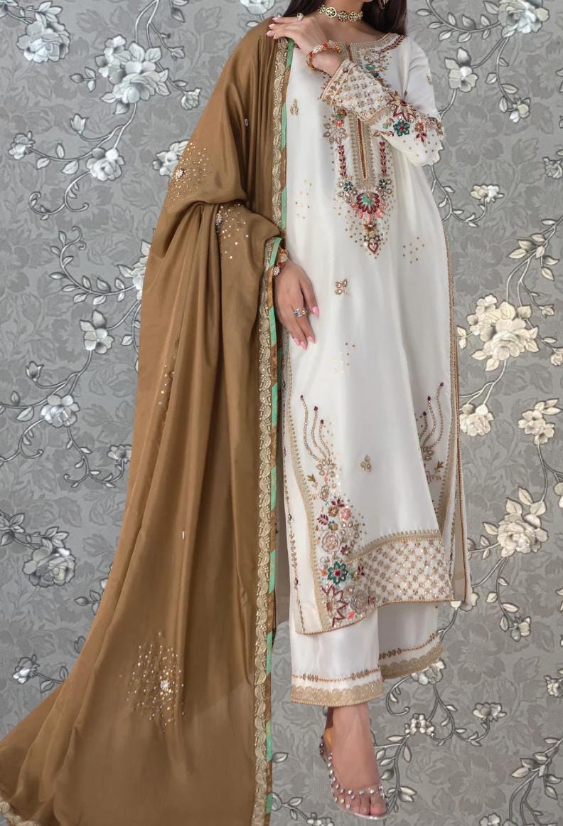 A woman wearing a white and gold dress with intricate embroidery, standing against a gray floral background.