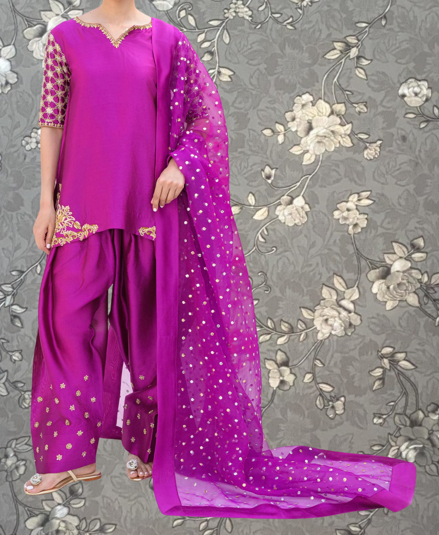 A person wearing a vibrant purple dress with gold embroidery and a matching purple shawl stands against a gray floral background.