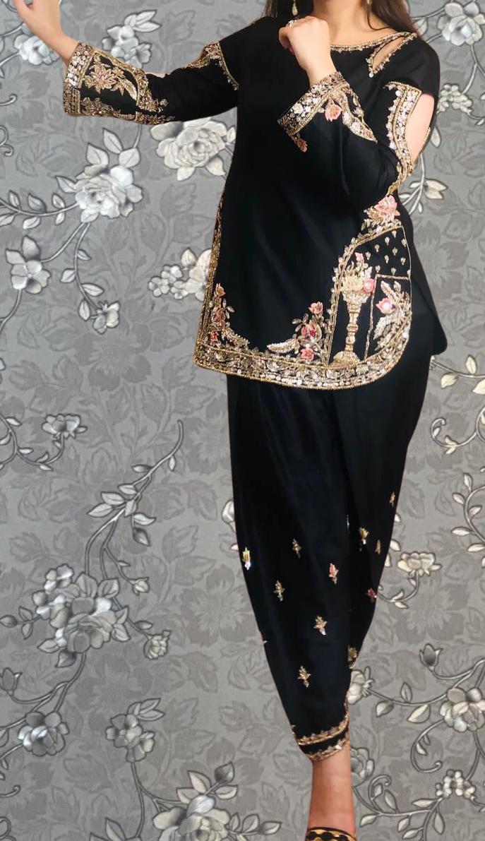 A woman wearing a black and gold dress with intricate embroidery poses against a gray floral background.