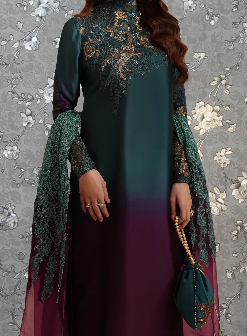 A woman wearing a long, green dress with intricate gold embroidery stands against a gray floral background.