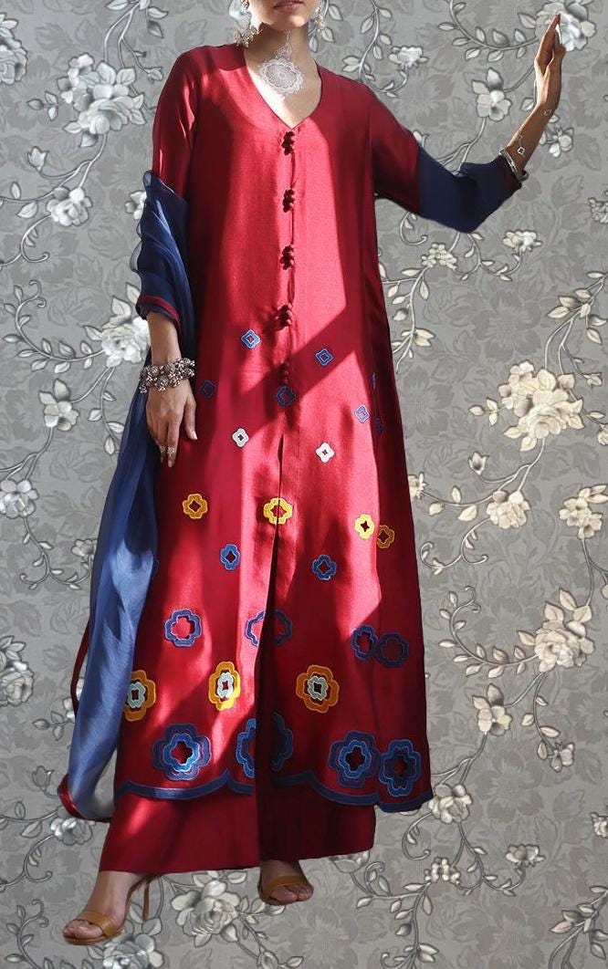A person wearing a long red dress with blue and yellow floral embroidery stands against a gray floral background.